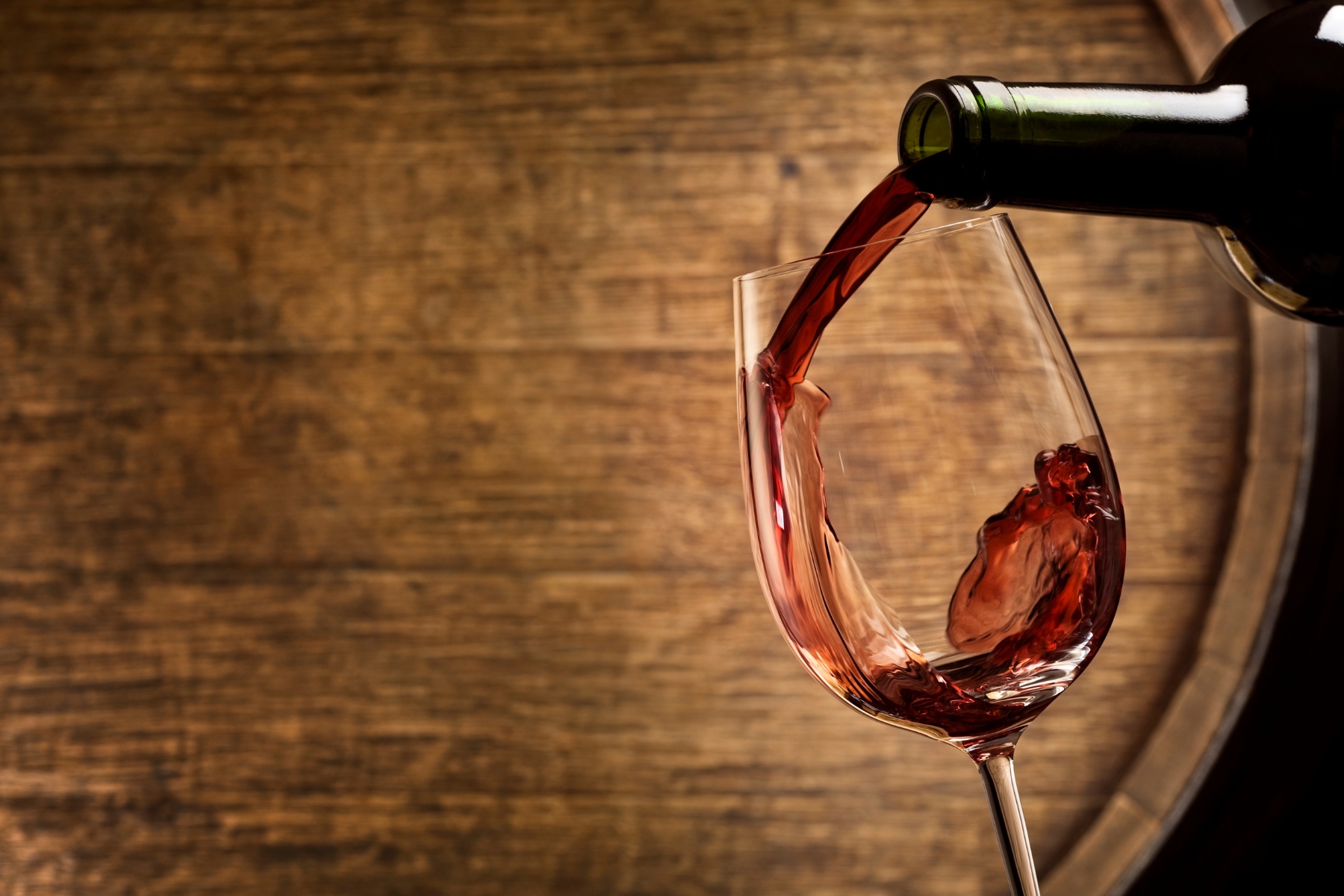Bottle of red wine being poured into a glass in front of a barrel